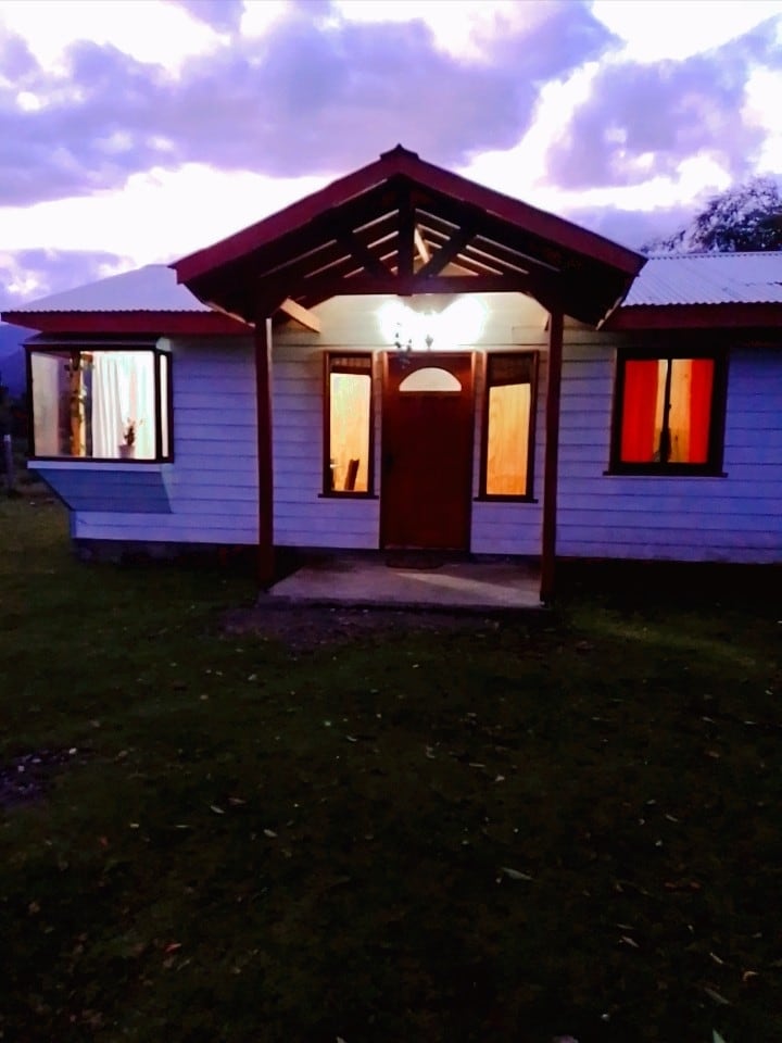 A charming cabin entrance is illuminated by a warm light, featuring a wooden door framed by large windows with soft curtains. The surrounding lawn is grassy, with a backdrop of cloudy twilight skies, creating a peaceful and inviting ambiance.