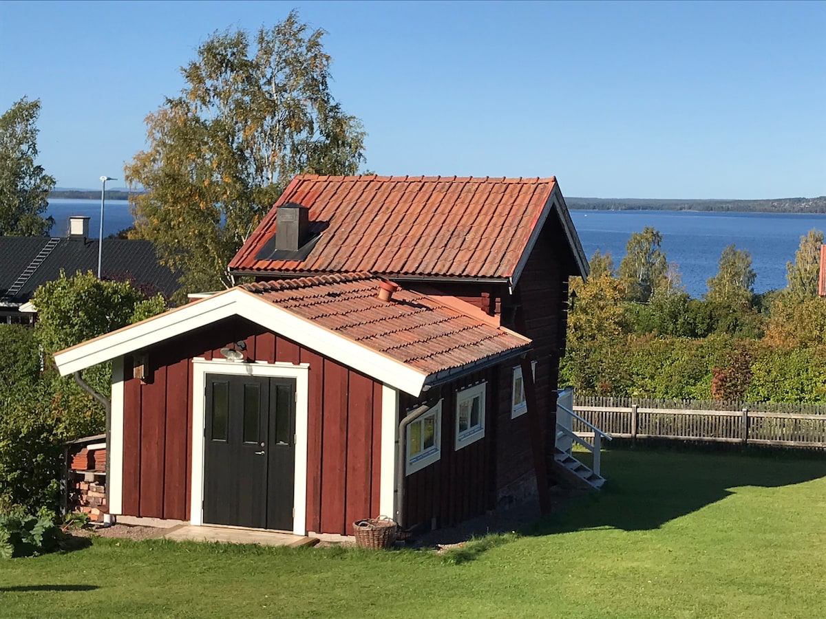 A charming wooden cottage is shown against a clear blue sky, featuring a red exterior with a sloped roof. A welcoming entrance with double doors faces a spacious lawn and offers views of the nearby lake and surrounding greenery.