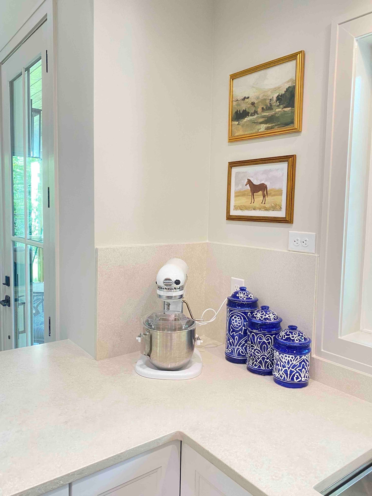 A modern kitchen corner is highlighted by a stand mixer on a light countertop. Decorative blue canisters are displayed next to a framed artwork featuring a horse. Natural light filters in from a nearby window, enhancing the serene atmosphere.