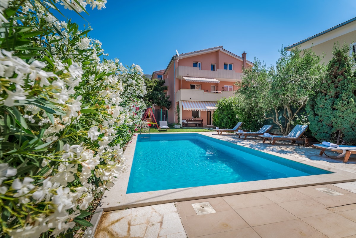 An outdoor swimming pool is surrounded by a paved area with sun loungers. Lush greenery and blooming flowers frame the space, providing a tranquil atmosphere. The villa's exterior, visible in the background, features a light-colored façade and offers access to the pool area.