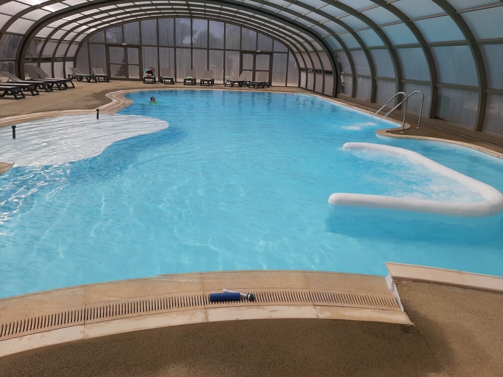 A heated, covered swimming pool is presented, featuring a clear blue water surface. The pool area includes a shallow section and a slide, with lounge chairs lined up along the perimeter. Natural light filters through the curved, transparent structure above.