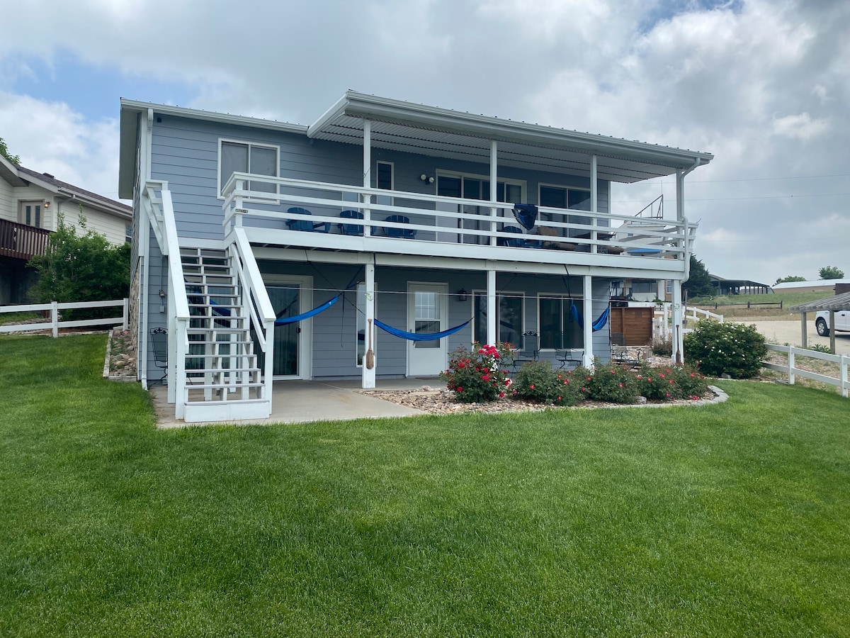 The exterior of a two-story home features light blue siding and a wide front porch adorned with comfortable chairs. A landscaped yard with green grass and flowering shrubs surrounds the house, while stairs lead to the upper level. The property includes a spacious deck with railings.