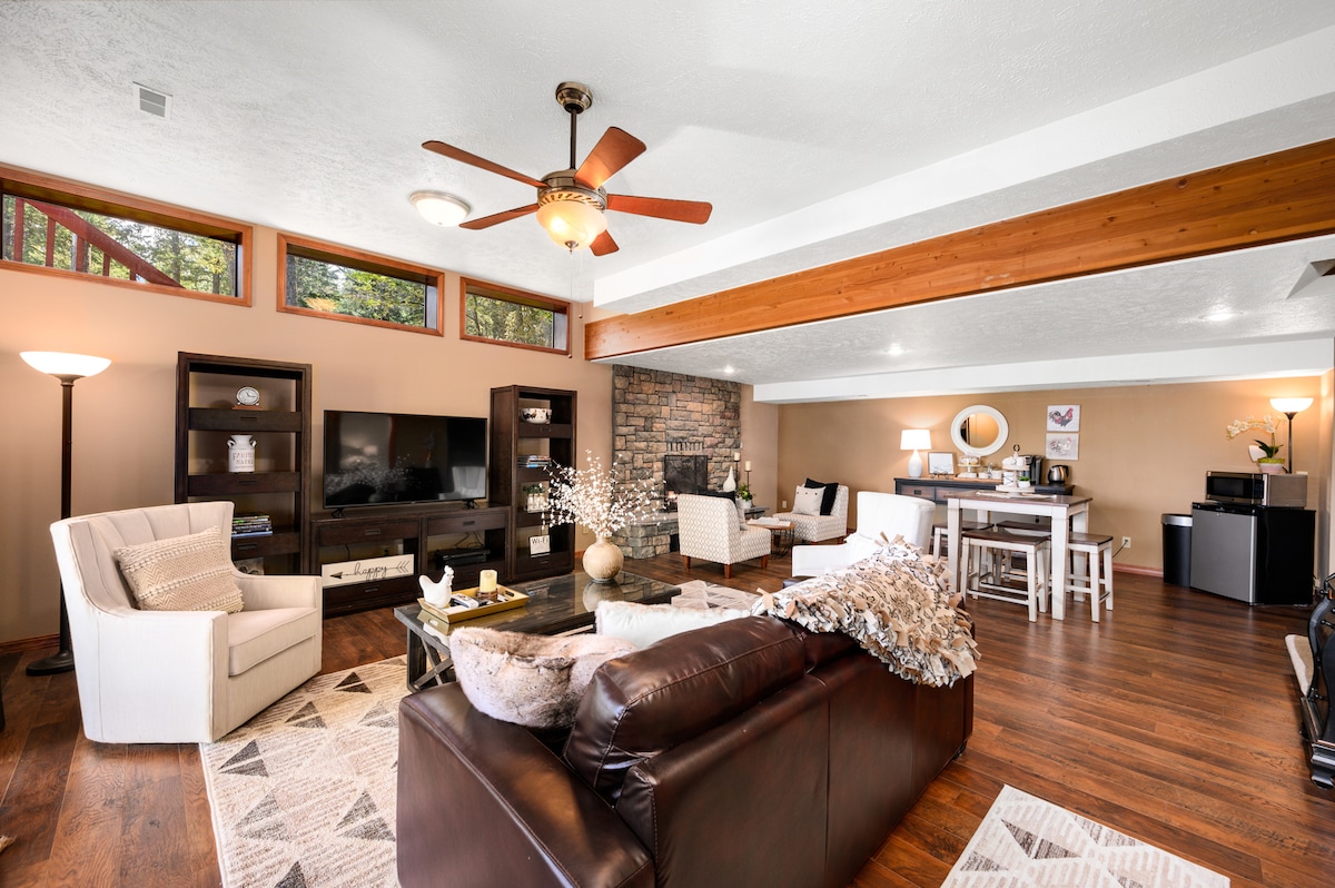 A spacious living area features a dark leather sofa, accompanied by a light armchair and a decorative coffee table. A stone fireplace is situated against a feature wall, and large windows allow natural light to fill the room. Additional furniture includes a dining table and a TV console.