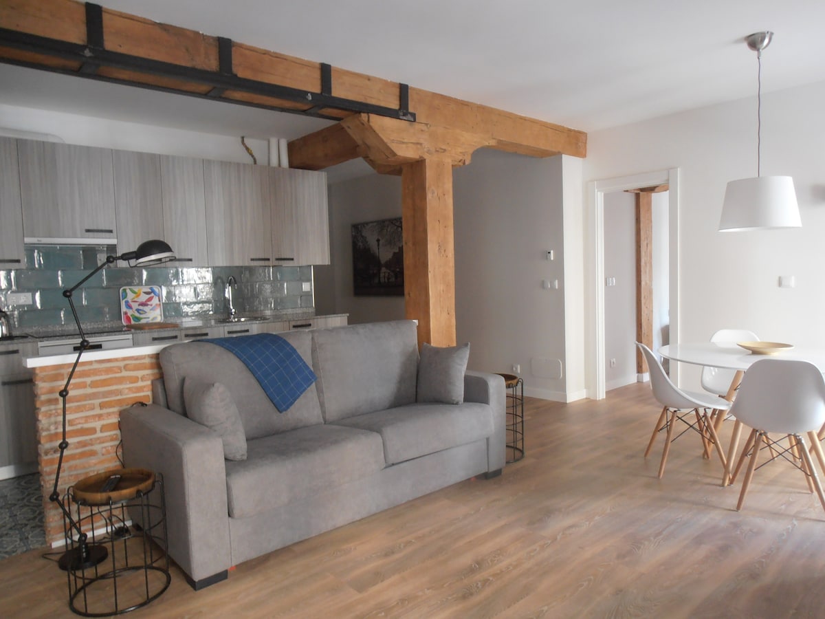 A spacious living area is presented, featuring a gray sofa with a blue throw, complemented by a modern dining table with white chairs. Exposed wooden beams and a brick wall provide a contrast to the contemporary kitchen equipped with sleek cabinets and tile backsplash.