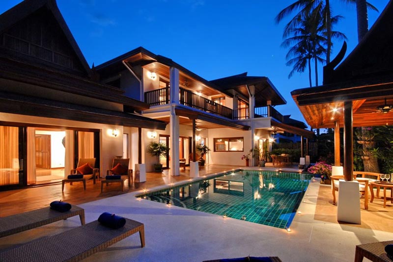 An inviting outdoor area showcases a blue-tiled swimming pool, surrounded by comfortable sun loungers. The villa's architectural features reflect traditional Balinese style, with warm lighting illuminating the exterior against a twilight sky. Lush greenery and palm trees frame the serene setting.
