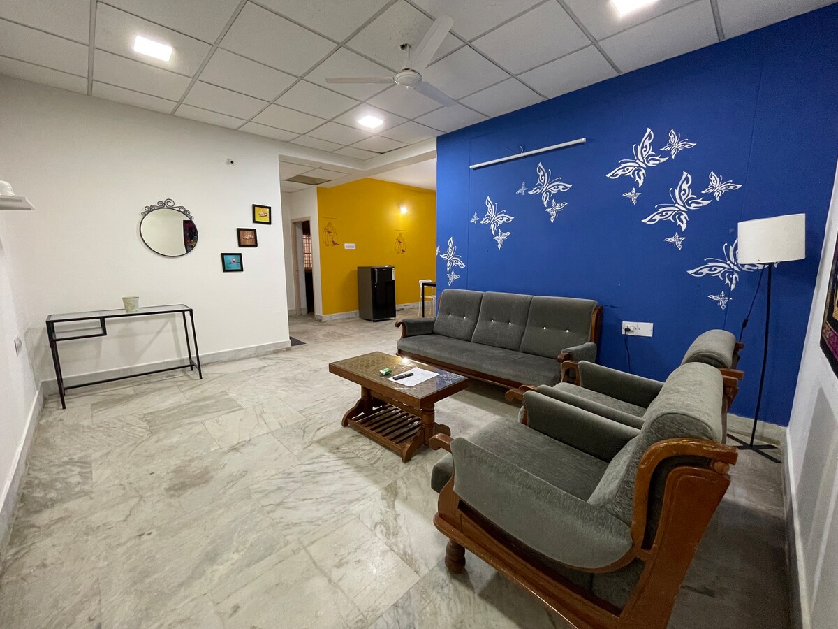The living area features a gray sofa and two cushioned chairs arranged around a wooden coffee table. A wall adorned with decorative butterflies in light blue contrasts with the bright yellow wall of the adjoining room. Natural light enters through the ceiling lights.