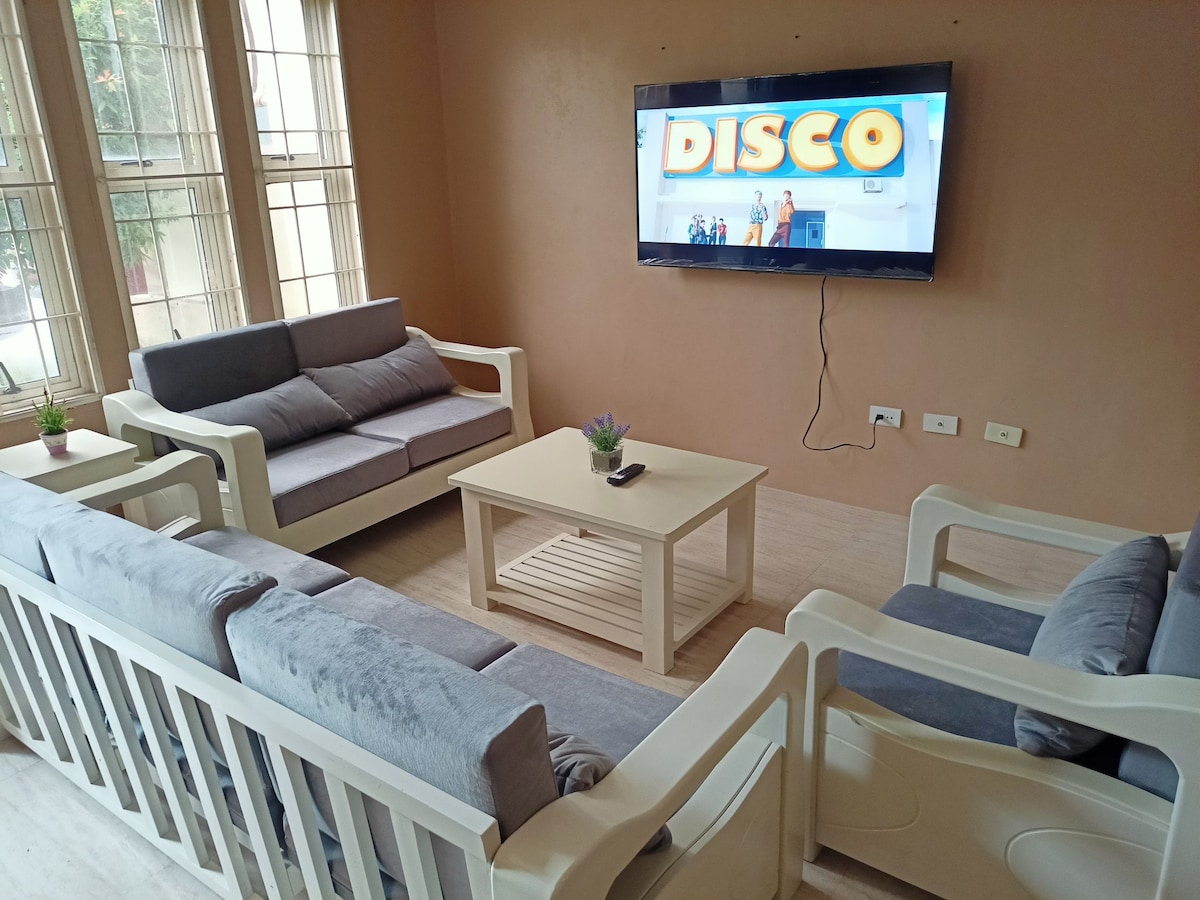 A comfortable seating area is arranged with light-colored sofas surrounding a low coffee table. A large television is mounted on the wall, displaying a vibrant screen. Natural light flows in through several windows, enhancing the space's cozy ambiance.