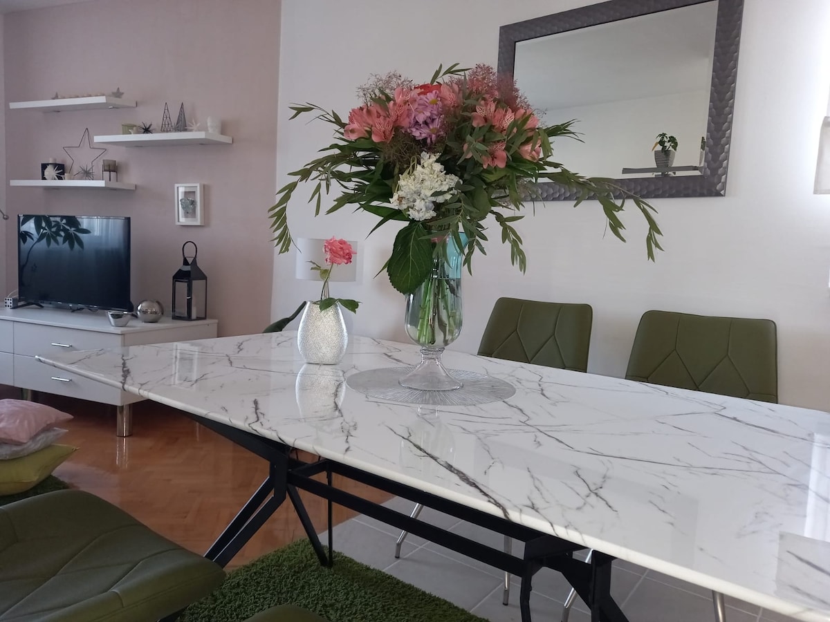 A modern dining area showcases a marble table complemented by green chairs. A large floral arrangement in a glass vase is the focal point, surrounded by a decorative small vase and a single pink flower. A mirror reflects light, enhancing the spacious ambiance.