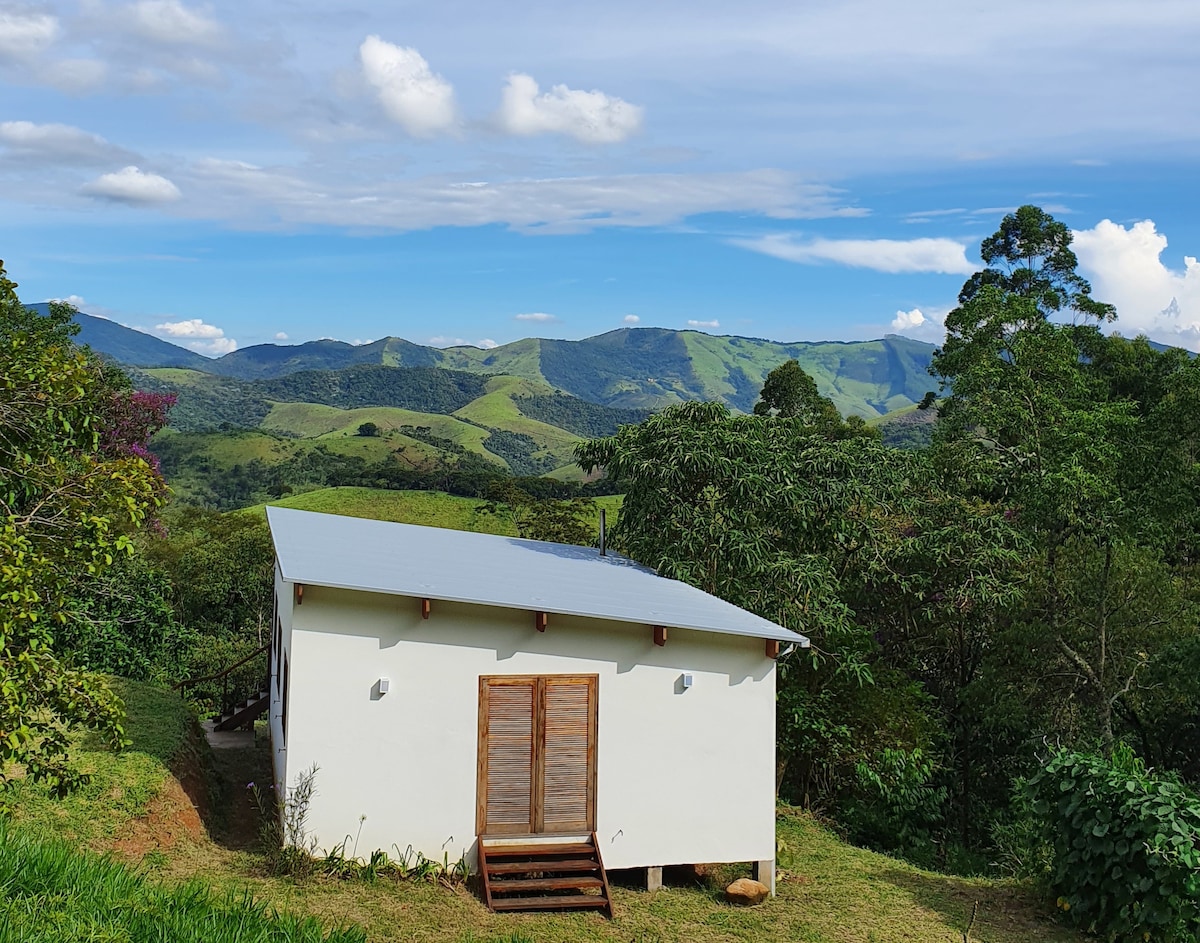 A charming house with a sloped roof is positioned on a hillside, surrounded by lush greenery. The back view reveals rolling mountains and a clear sky, providing a serene and scenic backdrop. A wooden porch with steps leads to the entrance.