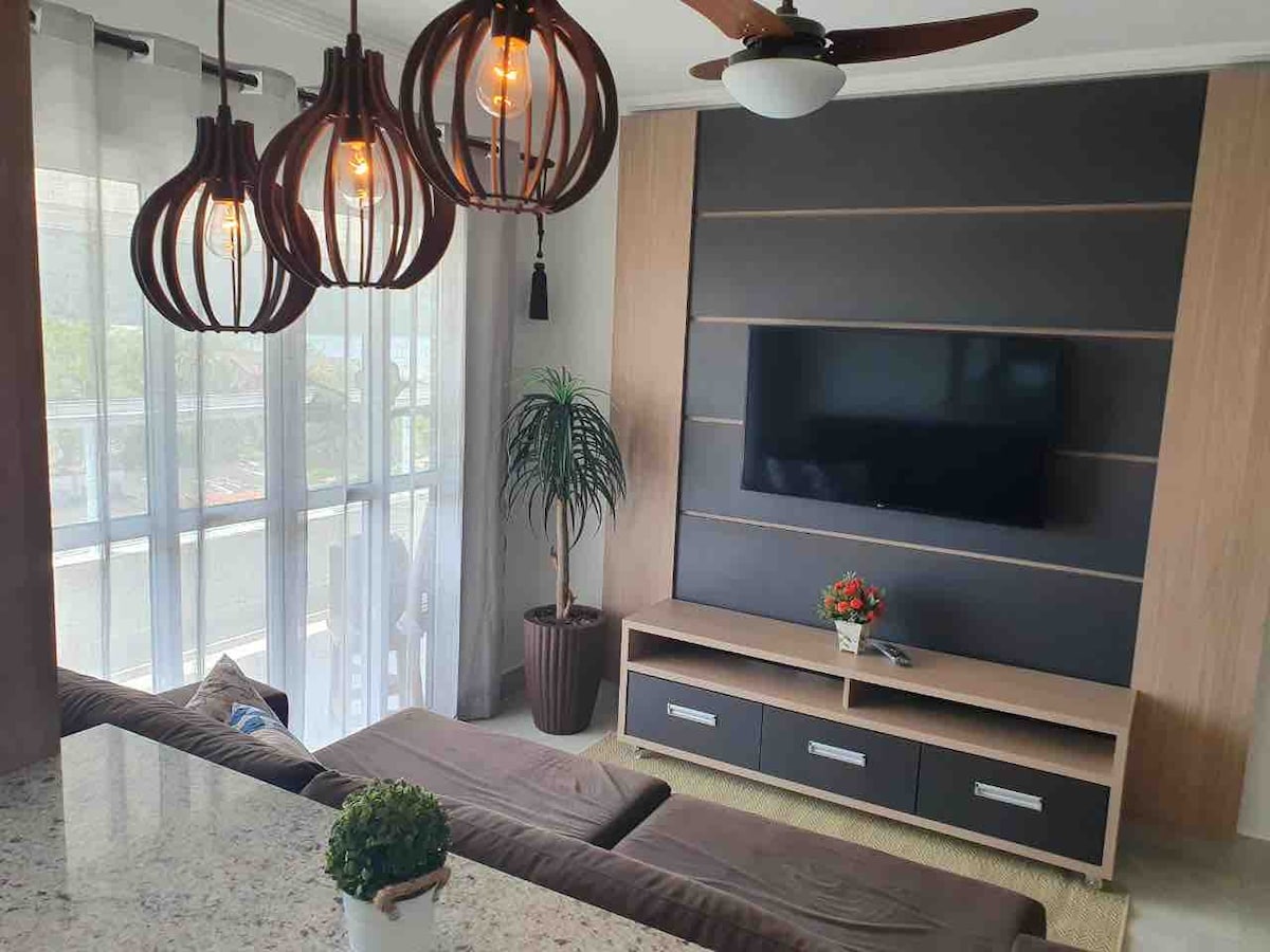 A cozy living area is presented with a modern design, featuring a comfortable brown sofa and a sleek TV unit. Three pendant lights hang from the ceiling, and a potted plant adds greenery. Large windows allow natural light to brighten the space.