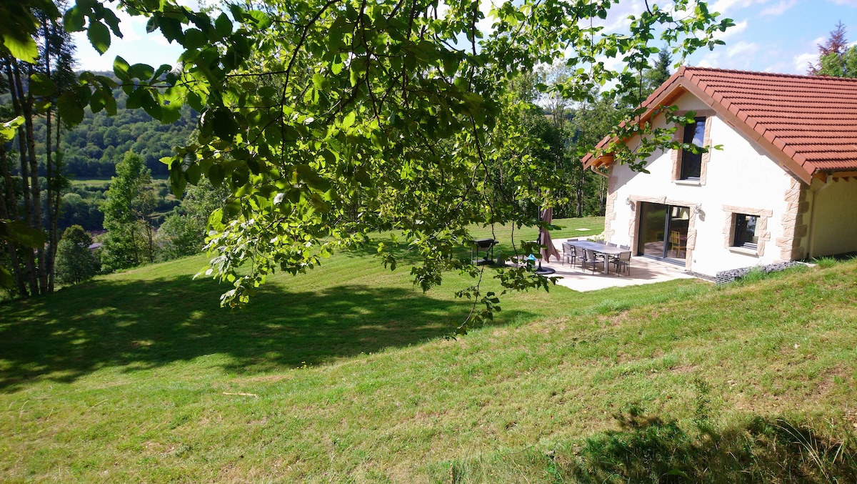 A welcoming gîte is set on a spacious green hillside, surrounded by trees. The exterior features large glass doors leading to an outdoor terrace, furnished with a dining table. The scenic view captures the surrounding natural landscape, enhancing the serene atmosphere.