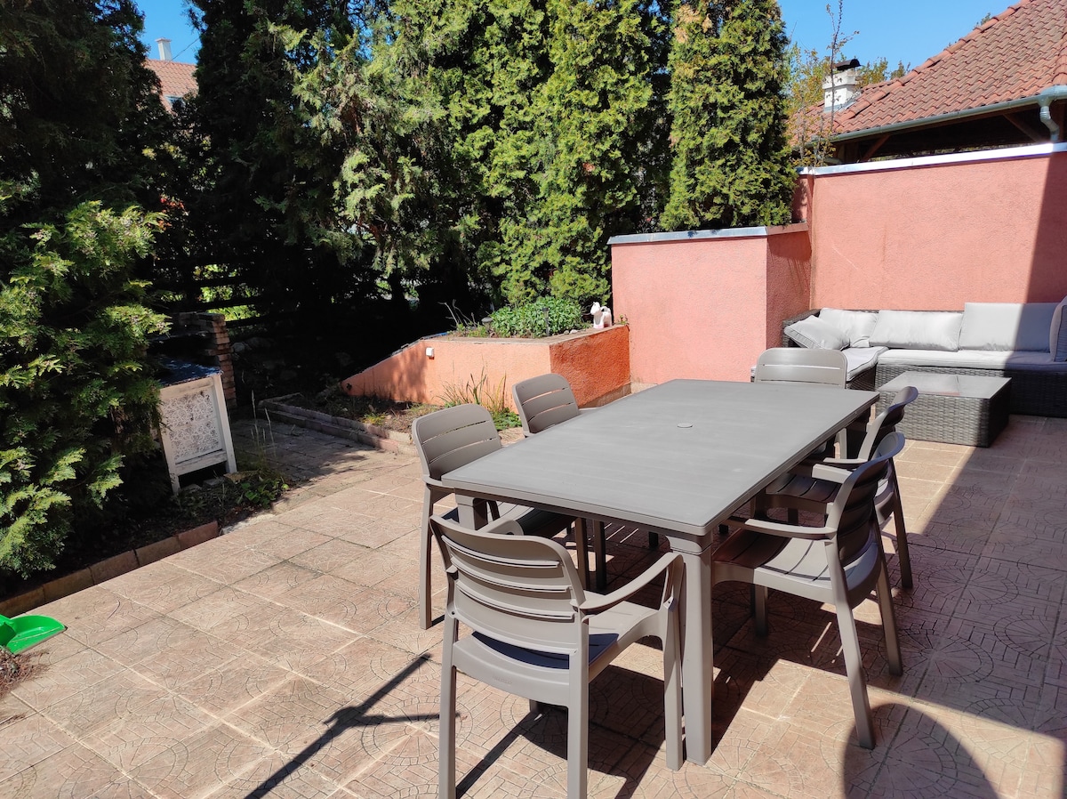An outdoor terrace is visible, featuring a large table surrounded by six chairs. A cozy seating area is situated against a wall, complemented by lush greenery in the background. The spacious area provides a suitable setting for gatherings or relaxation.