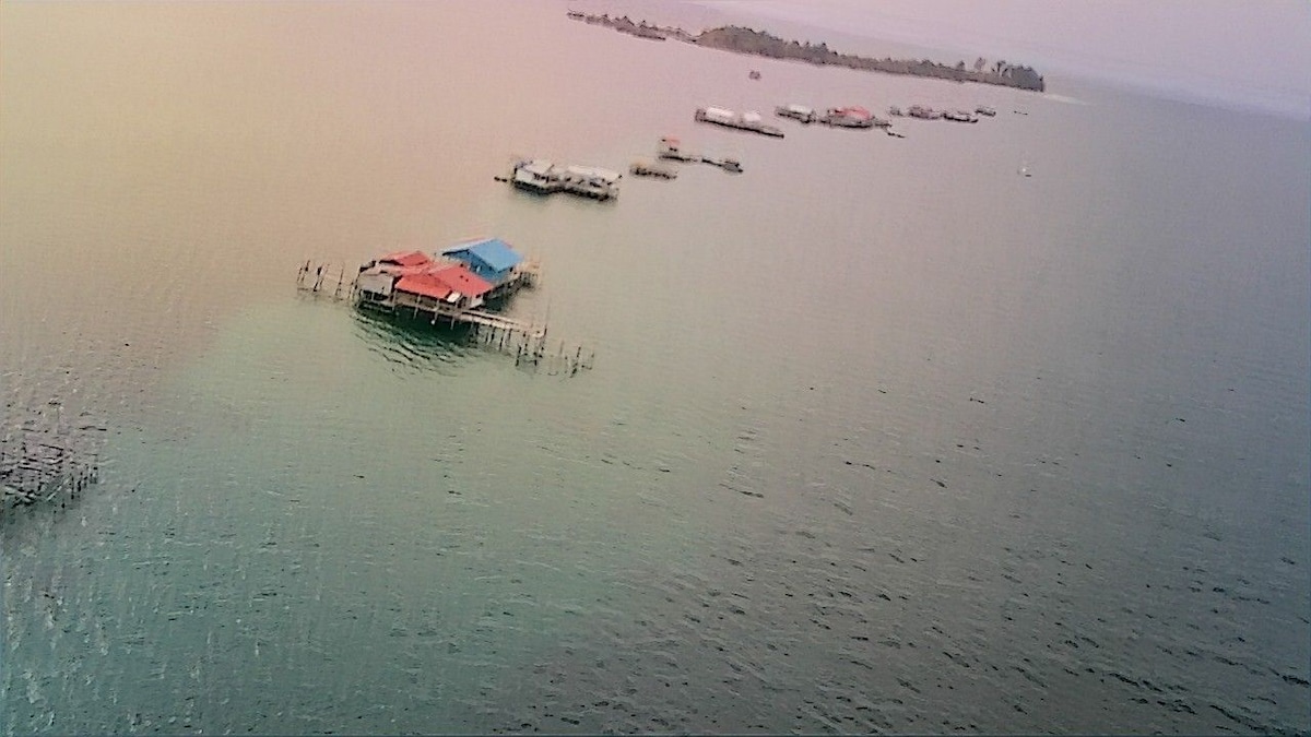 A collection of stilted structures is visible on the calm waters, showcasing a variety of roofs in shades of blue and red. The surrounding ocean reflects soft hues, creating a serene maritime ambiance. Nearby islands can be faintly seen in the distance.