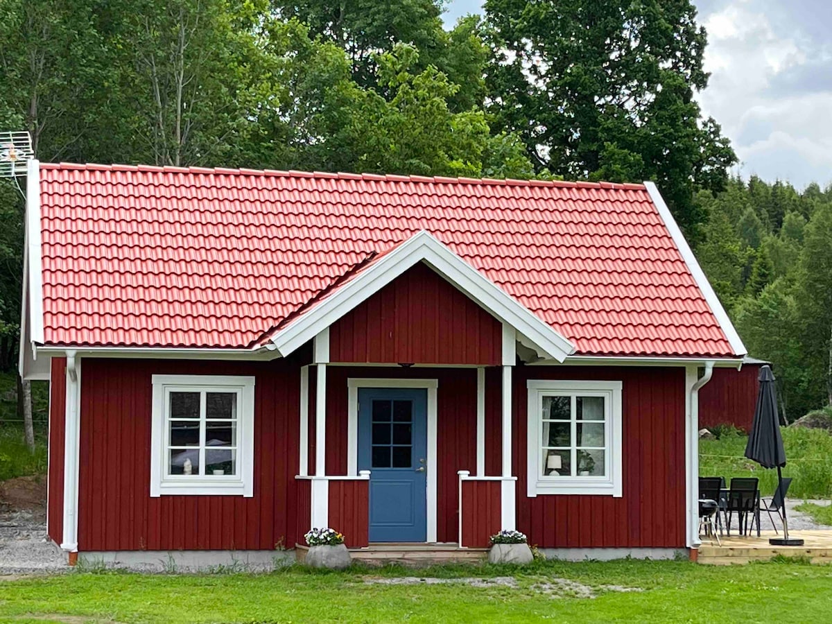 A charming red house with a bright blue front door is surrounded by lush greenery. White window frames highlight the two front windows, and a spacious outdoor seating area is visible on the right. The sloping roof is covered with red tiles, contributing to the inviting exterior.