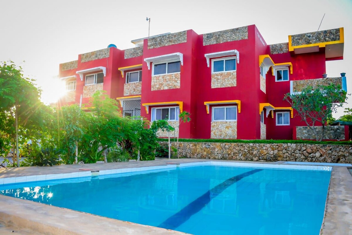 A vibrant red building featuring a stone accent exterior is presented. The structure includes several windows with yellow trim. A clear swimming pool is located in the foreground, reflecting sunlight, while landscaped greenery surrounds the area.