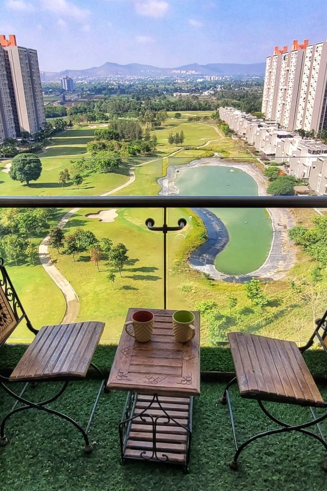 A private terrace showcases two wooden chairs paired with a small table, inviting relaxation. Beyond, sweeping views of the lush golf course and tranquil water feature extend to the distant hills under a clear blue sky.
