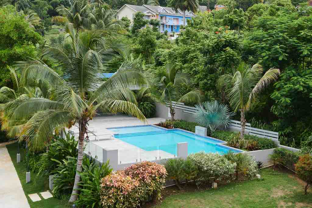 A serene outdoor area features a clean, blue swimming pool surrounded by lush greenery and palm trees. Soft landscaping provides a sense of privacy, while the pool area is paved with light-colored tiles, creating a calm and inviting space.