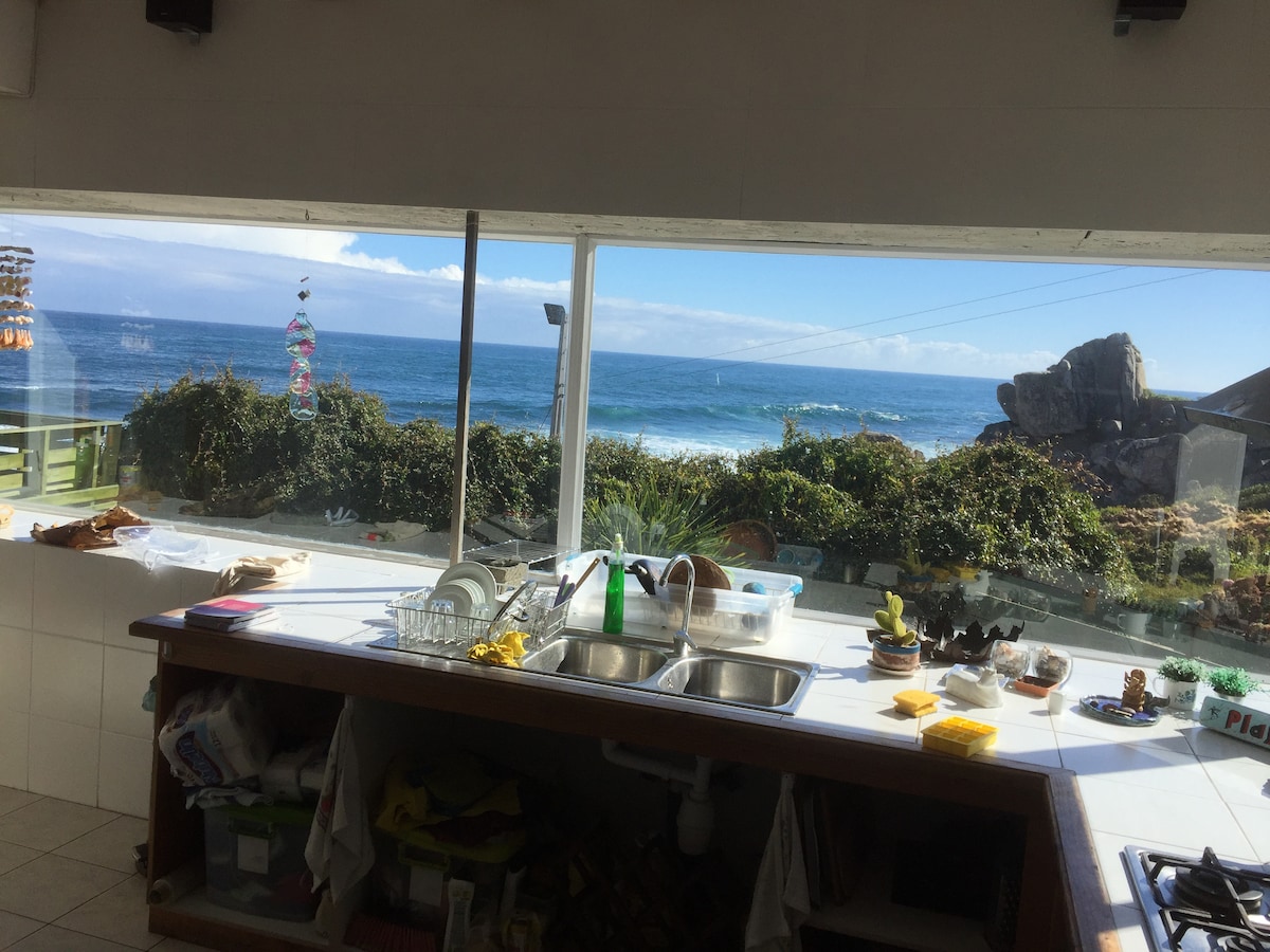 A well-equipped kitchen features a central island with a dual sink and a view of the ocean through expansive windows. Natural light illuminates the space, highlighting a variety of kitchen supplies, and plants are visible outside the window near the rocky shoreline.