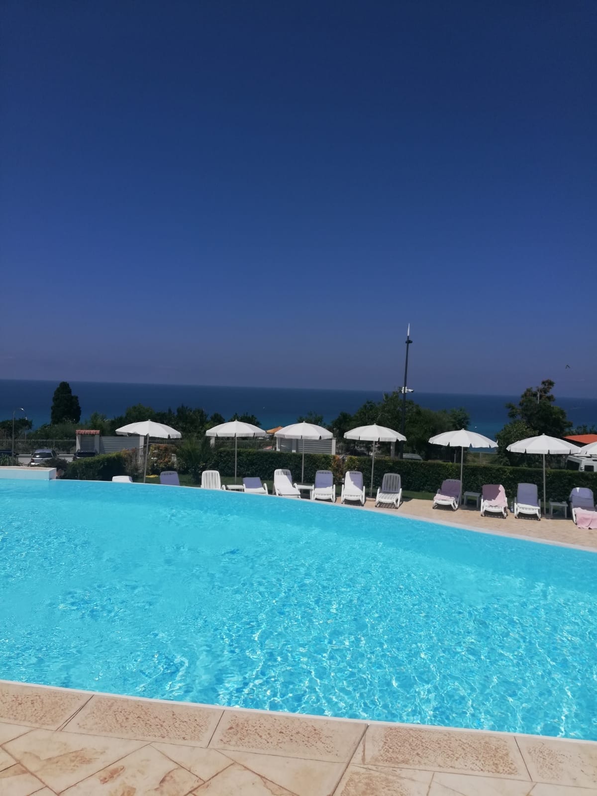 A large swimming pool features crystal-clear water, surrounded by comfortable lounge chairs. Beach umbrellas provide shade for relaxation. The distant coastline is visible, offering a serene view of the sea under a clear blue sky.