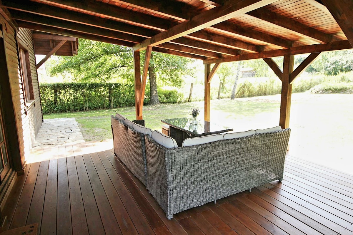 A spacious covered terrace features a comfortable seating arrangement with plush cushions and a coffee table. The wooden beams and flooring enhance the natural feel, while a garden is visible in the background, providing a serene outdoor setting.