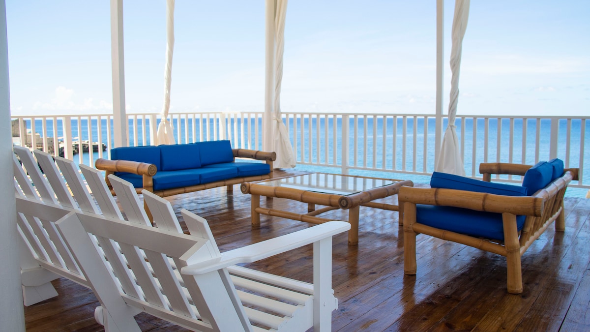 A spacious outdoor lounge area is furnished with bamboo chairs and a glass-top table, offering a relaxing view of the sea. Blue cushions add comfort to the seating arrangement, which is set against a backdrop of clear skies and gentle waves.