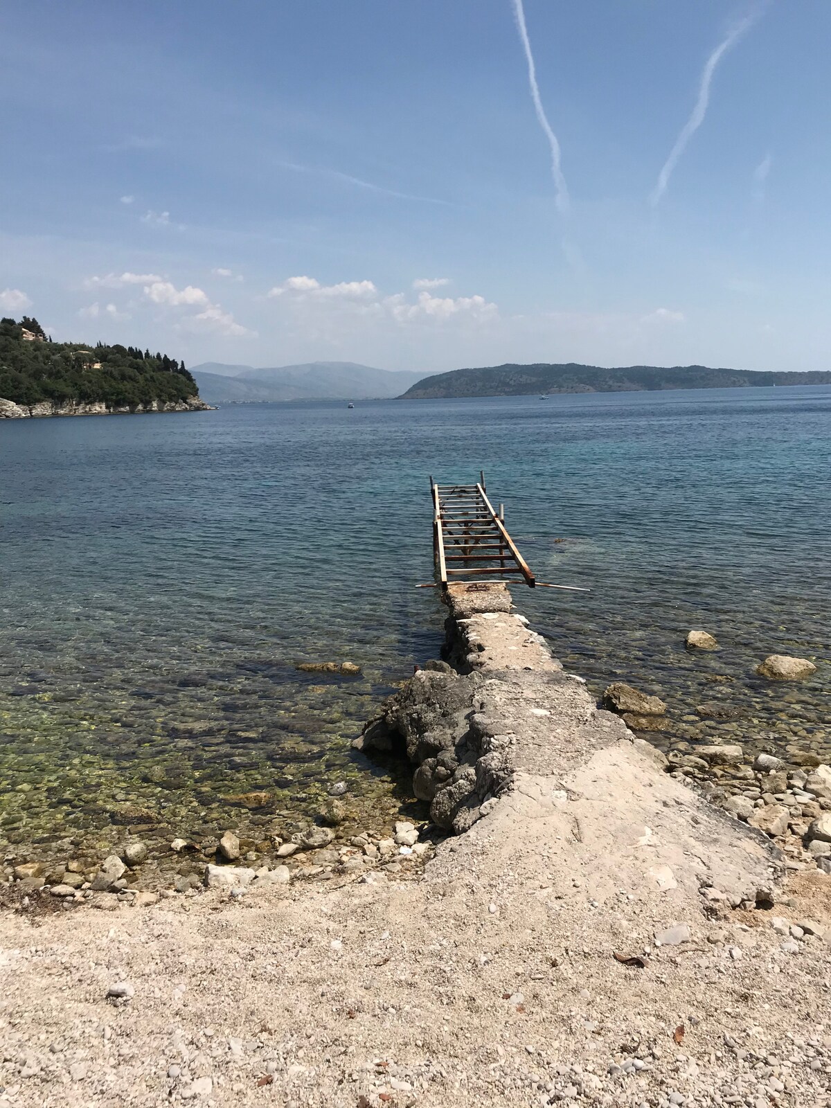 A sturdy stone pier extends into calm waters, leading towards the horizon. The clear water reveals a pebbly shoreline, with gentle waves lapping against the edge. Lush hills frame the background under a clear blue sky, enhancing the serene coastal view.