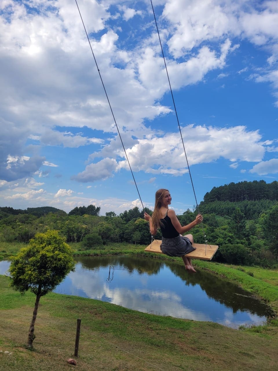 A swing is positioned against a backdrop of vibrant greenery and a tranquil pond. The sky is adorned with scattered clouds, creating a serene outdoor setting. Nearby, a tree stands, adding to the natural ambiance of the location.
