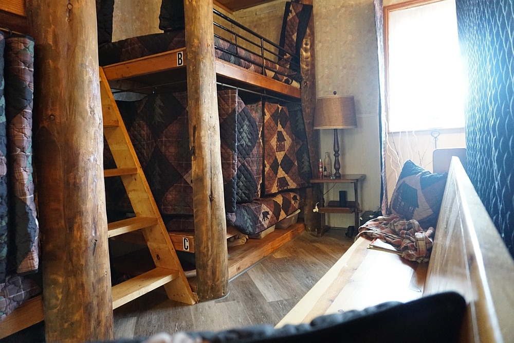 A shared sleeping area features built-in wooden bunks nestled against rustic wooden posts. Bunk B is positioned above another bunk, with privacy curtains drawn. Quilted bedding in earthy tones adds warmth. A small bedside lamp and a soft seating area provides functional space within the room.