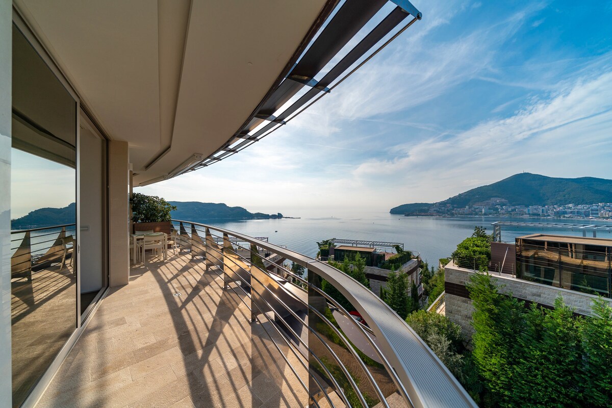 A spacious terrace features a curved railing, offering unobstructed views of the Adriatic Sea and surrounding hills. Sunlight casts shadows on the polished stone floor, and seating arrangements are visible, inviting relaxation amidst the scenic backdrop.