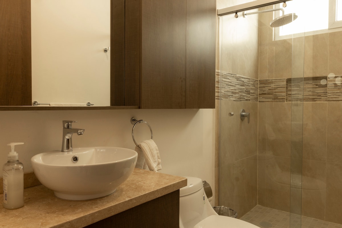 A modern bathroom features a glass-enclosed shower with tiled details, alongside a sleek sink set upon a stone countertop. Neutral-toned cabinetry complements the design, while natural light filters through a small window.