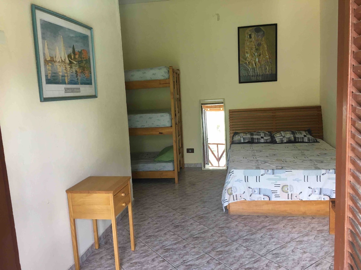 A well-lit room features a spacious bed with a patterned bedspread, located adjacent to a wooden bunk bed. A small wooden table is positioned beside the bed, and a mirror reflects the room. Light colored walls enhance the open feel of the space.