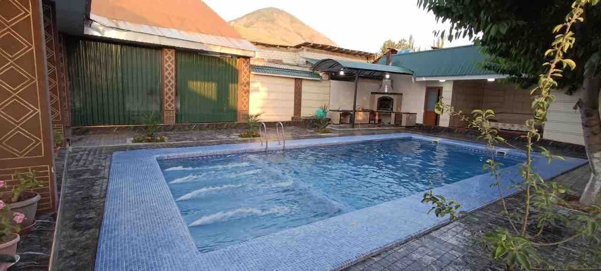 A serene swimming pool is surrounded by a tiled patio, complete with lounge areas. Overhead, the backdrop features a mix of covered and open spaces, with a barbecue area visible. Lush greenery frames the setting, enhancing the inviting outdoor space.