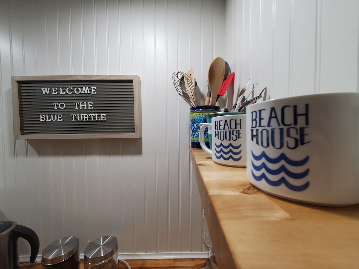 The kitchenette features a wooden shelf displaying two beach-themed mugs and additional cooking utensils. A letter board nearby welcomes guests to 'The Blue Turtle,' creating a friendly and inviting atmosphere. Stainless steel canisters are visible on the countertop.