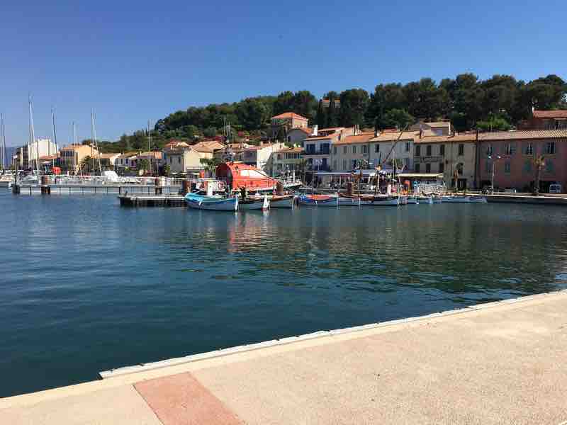 The image features a serene waterfront scene with colorful boats anchored in calm waters. A charming village with pastel buildings is visible in the background, surrounded by lush greenery. The clear blue sky creates a bright atmosphere, reflecting off the water's surface.