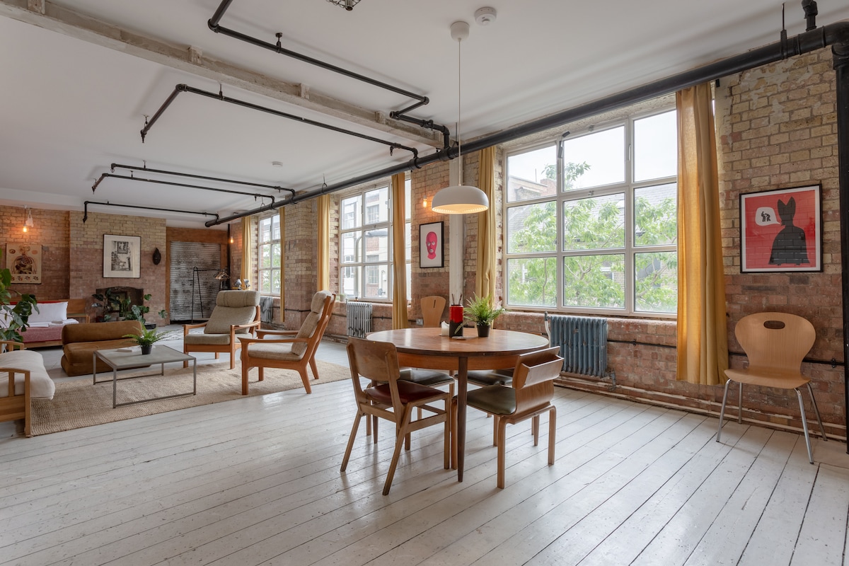 An open-plan living area features large windows allowing natural light to illuminate the space. A circular dining table is surrounded by wooden chairs, while a cozy seating arrangement includes a sofa and armchair. Contemporary artwork decorates the brick walls, adding character to the loft.