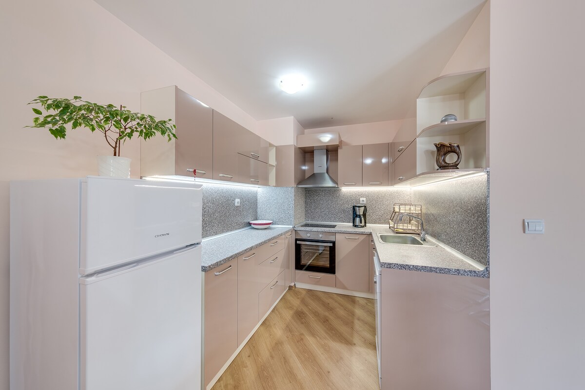 A modern kitchen features sleek cabinetry in light tones, complemented by a gray countertop. Essential appliances, including a refrigerator and oven, are neatly integrated. Soft lighting highlights the workspace, while a small plant adds a touch of greenery to the room.