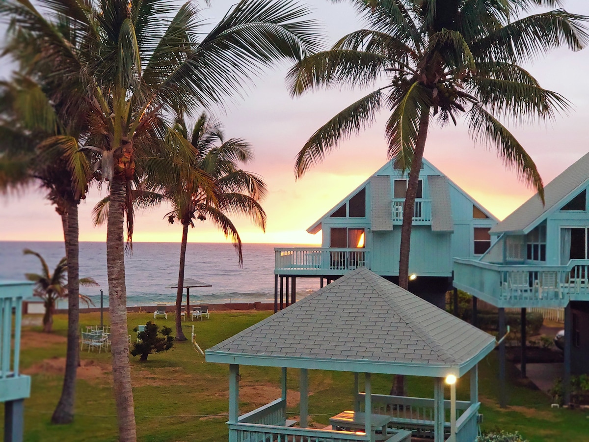A view of colorful sunset sky is framed by tall palm trees above the ocean. Two villas with gables are positioned among lush grass, complemented by a gazebo in the foreground. Tables and chairs are visible off to the side, suggesting a relaxing outdoor space.