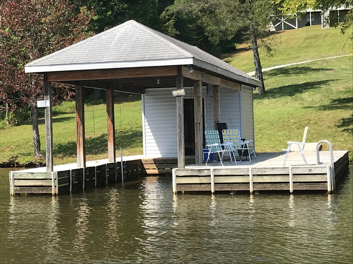 A boathouse is situated at the edge of the lake, featuring a covered area and a spacious deck. Several outdoor chairs are arranged neatly for seating. The gentle slope of the grassy yard leads up to the property, surrounded by trees and natural greenery.
