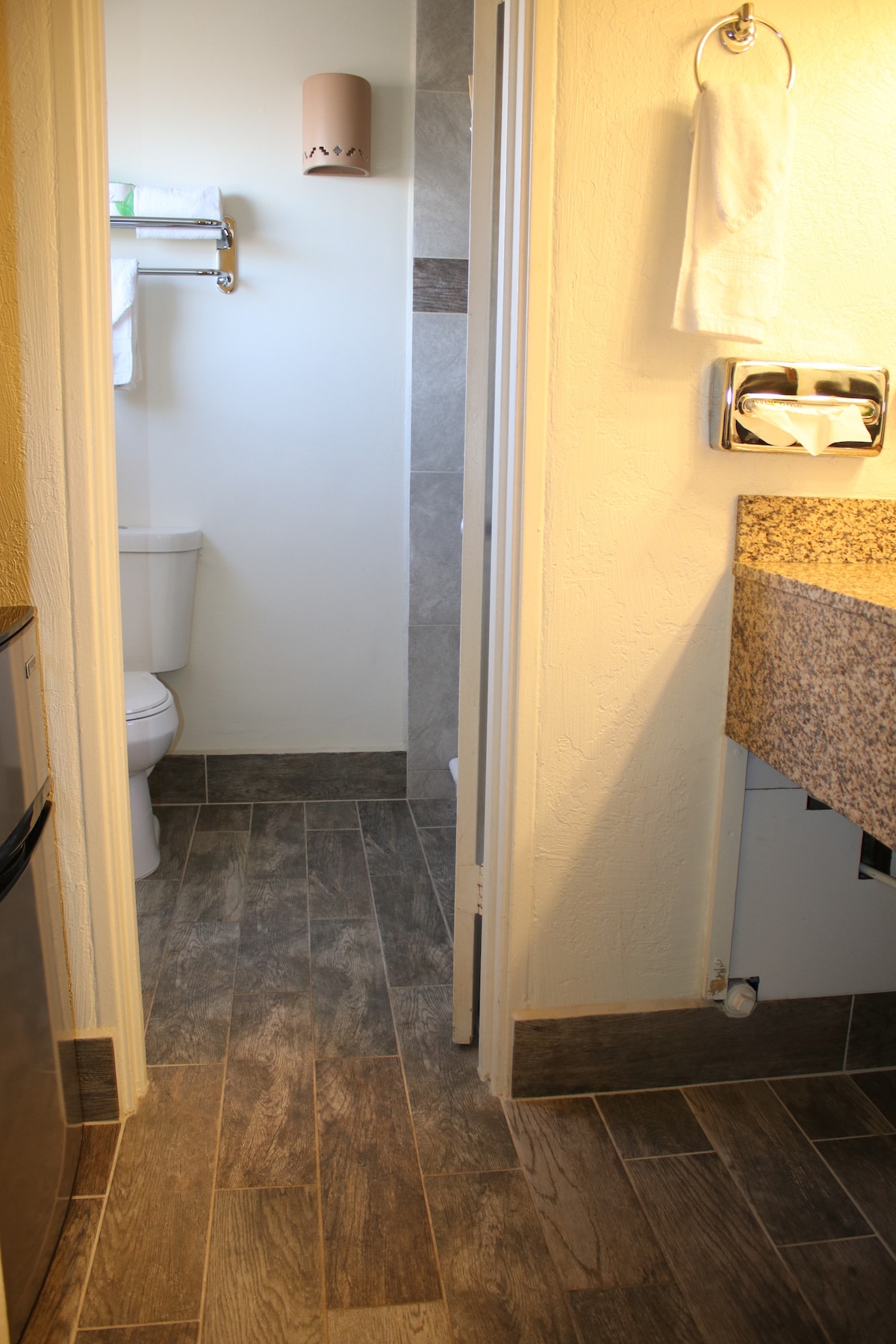 A bathroom area is seen, featuring a toilet and a granite countertop with a sink. Tile flooring adds a modern touch, while a small shelf holds towels. A shower is visible in a corner, complete with gray tiling. A compact refrigerator can be seen at the entrance.