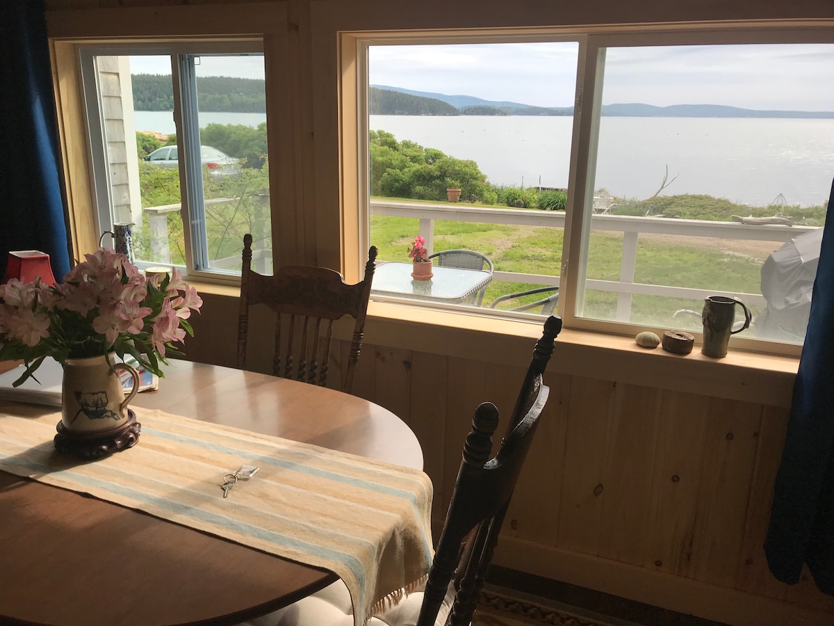 A dining area is visible featuring a wooden table set with a light table runner. A vase of fresh flowers adds a pop of color. Large windows frame a view of Frenchman Bay, with greenery and a distant shoreline outside.