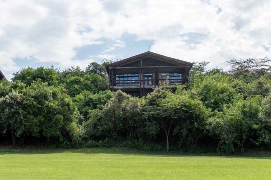 A cottage is nestled among lush greenery, showcasing large windows that invite natural light inside. The landscaping features tall grass, enhancing the serene setting. The structure's pitched roof and wooden façade provide a rustic charm, surrounded by the vibrant foliage of the golf course landscape.