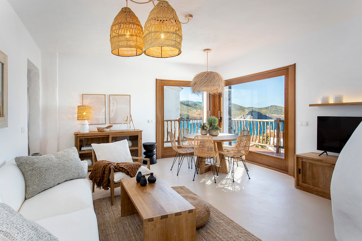 A bright living area features natural materials and textures, with a spacious sofa and wooden coffee table. Large windows provide a scenic view of the surrounding landscape and invite ample light. Elegant pendant lights hang from the ceiling above a dining table set for six.