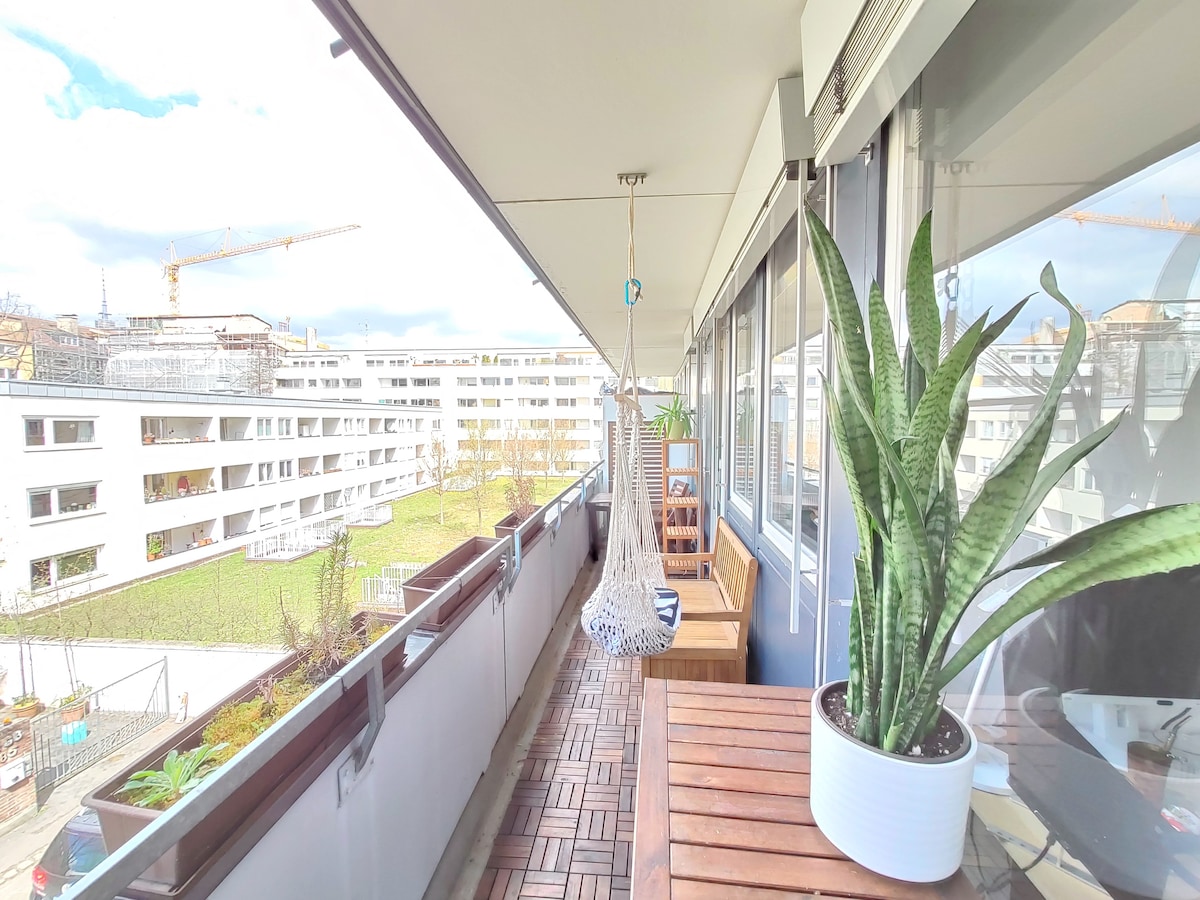 A spacious balcony is depicted, featuring a hanging chair and potted plants. The wooden flooring adds warmth to the area, while the view showcases a green space and buildings in the distance. Natural light brightens the outdoor space, creating an inviting atmosphere.