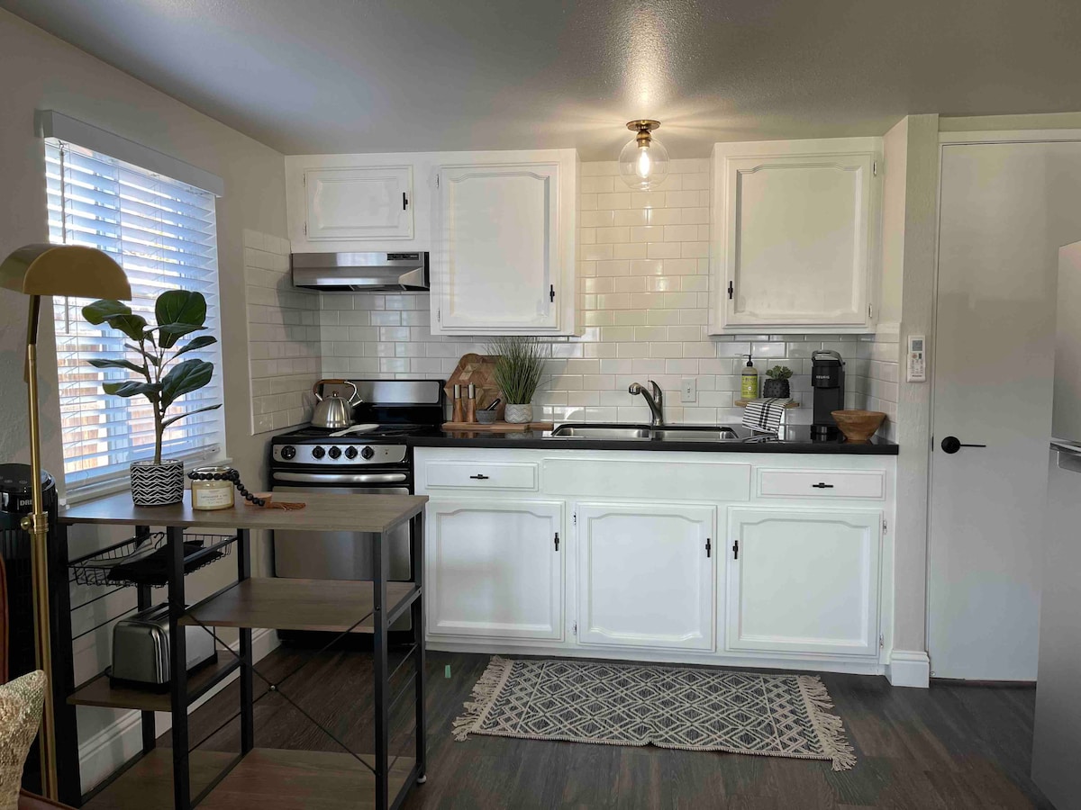 A functional kitchen area is presented, featuring white cabinetry and black countertops. Stainless steel appliances, including a gas stove and refrigerator, are visible. A small table with a decorative plant adds a touch of greenery, while natural light filters in through the window with white blinds.