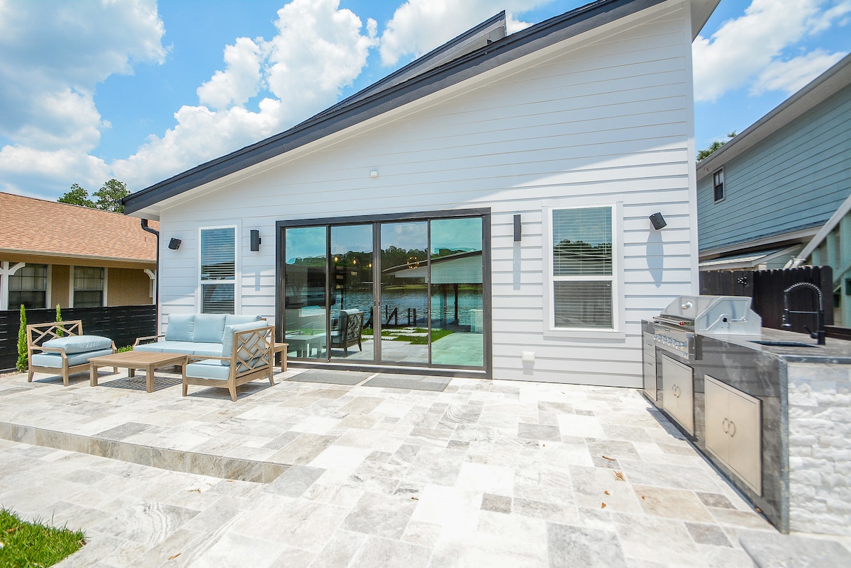 The exterior of the house is shown, featuring a spacious outdoor area with gray stone flooring. A comfortable seating arrangement is positioned near large sliding glass doors that open to a lakefront view. A propane grill is visible at the side, enhancing outdoor dining options.