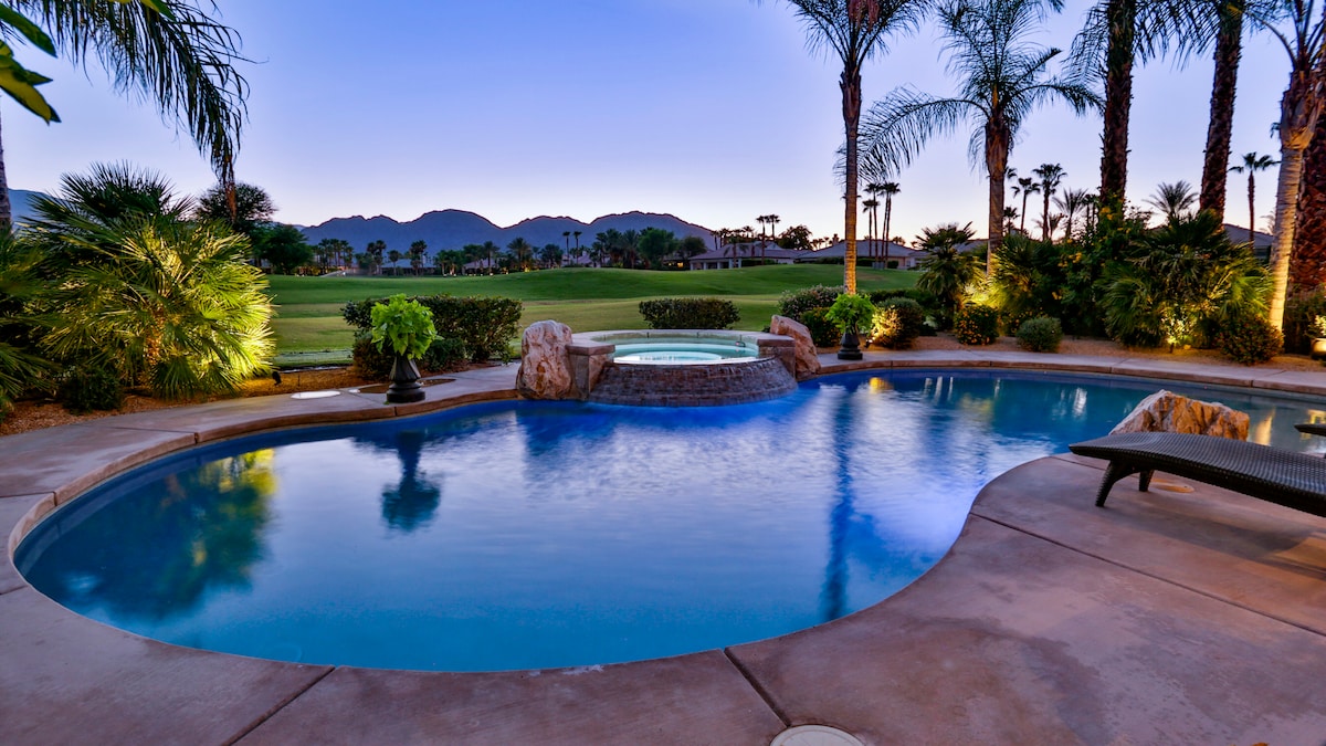 A serene saltwater pool is framed by lush palm trees and mountain views. Soft lighting highlights the water's surface, while a spa is positioned beside the pool. Natural stone accents are visible, adding texture to the outdoor space.