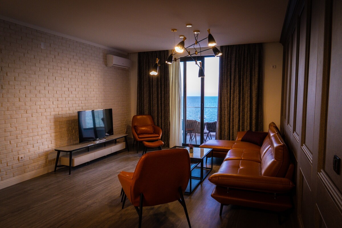A well-lit living area features large windows with views of the sea. A modern television sits on a low table across from a spacious sectional sofa and two accent chairs. Bright curtains frame the windows, highlighting the inviting ambiance of the space.