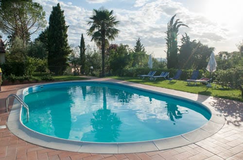 A rounded swimming pool is surrounded by a tile deck and lush greenery. Sun loungers are arranged nearby, and tall trees provide shade. The sky is partly cloudy, and reflections are visible on the pool's surface, enhancing the tranquil setting.