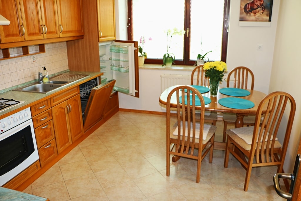 A functional kitchen space features a wooden cabinetry setup, equipped with a gas stove, oven, and refrigerator. A round dining table with four wooden chairs is set with turquoise placemats. Natural light streams through a window adorned with potted plants.