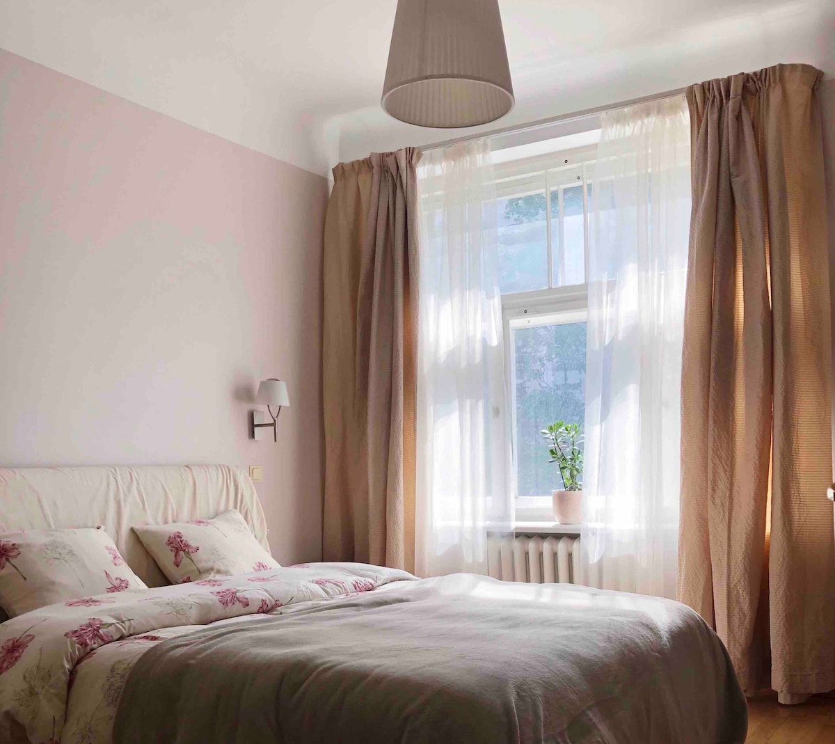 A bright bedroom is presented with soft pink walls and large windows adorned with sheer curtains. A spacious bed features floral-patterned linens, while a small potted plant sits on the windowsill, adding a touch of greenery to the serene atmosphere.